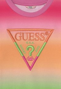 T-shirt w odcieniach różu i pomarańczy, z haftowanym logo w kształcie trójkąta z napisem "GUESS®" oraz "WASHED JEANS" w żywych kolorach.