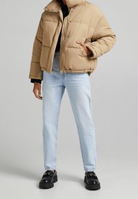 Giacca puffer beige con colletto alto, chiusura con zip e tasca con pattina; indossata sopra un maglione nero e jeans azzurri chiari; abbinata a scarpe nere.