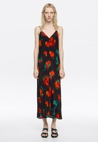 Junge Frau trägt ein schwarzes ärmelloses Kleid mit roten und grünen Blumenmustern und Riemchensandalen, steht vor einem weißen Hintergrund.