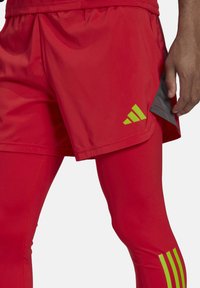 Rode sportieve shorts hebben een lichtgewicht ontwerp met een zijventilatie, gecombineerd met aansluitende rode leggings. Groene merktekens zijn zichtbaar.