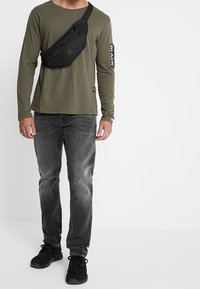 Svart axelväska, olivgrön långärmad skjorta och bleka grå jeans. Sneakers är svarta med texturerad detaljering. Armen har en vit tryckt etikett.