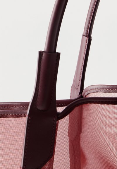 Κοντινό πλάνο χειρολαβών τσάντας από μπορντό δέρμα ραμμένων σε ημιδιαφανές ροζ ύφασμα, με λευκό φόντο.