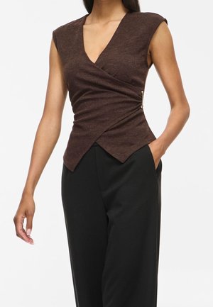 Femme portant un haut cache-cœur sans manches marron foncé avec une fermeture latérale et un pantalon noir taille haute, une main dans la poche.
