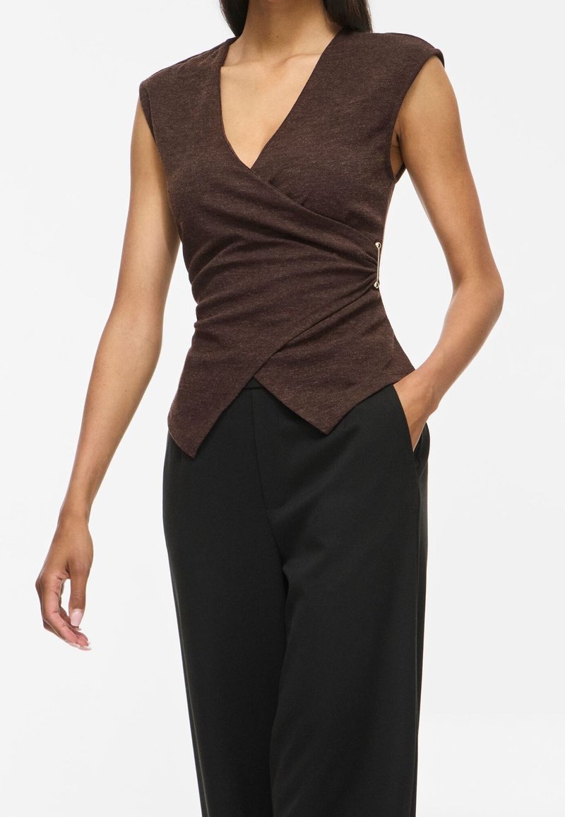 Femme portant un haut cache-cœur sans manches marron foncé avec une fermeture latérale et un pantalon noir taille haute, une main dans la poche.