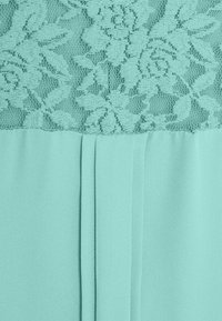 Um vestido verde menta com um sobreposição de renda com padrões florais na parte superior e um tecido liso e plissado na parte inferior.