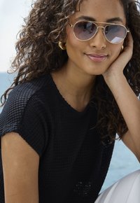 Schwarzes strukturiertes Strickoberteil, hellfarbige Sonnenbrille mit goldenem Rahmen und große goldene Creolen. Der Hintergrund zeigt Wasser und Himmel.