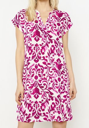 Femme portant une robe blanche à manches courtes avec un motif floral et tourbillonnant rose vif et un décolleté en encoche.