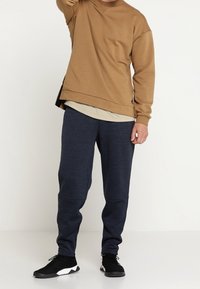 Sudadera marrón con cuello redondo, superpuesta a una camisa beige. Combinada con pantalones deportivos azul oscuro y zapatillas deportivas negras.