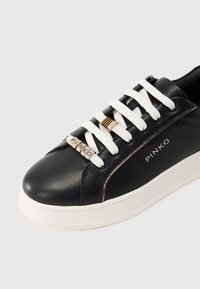 Μαύρο δερμάτινο αθλητικό παπούτσι με λευκά κορδόνια, μεταλλικά στοιχεία σε χρυσό τόνο και υφή φινιρίσματος. Διαθέτει τη σήμανση "PINKO" στο πλάι και λευκή σόλα.