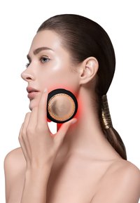 Svart och guld handhållen enhet med en texturerad cirkulär yta som hålls nära en persons käklinje; avger rött ljus för hudvårdsändamål.