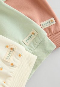 Tre articoli di abbigliamento in morbidi colori pastello: rosa polveroso, verde chiaro e crema, caratterizzati da motivi floreali e etichette con la scritta "FELICE E LUMINOSO".