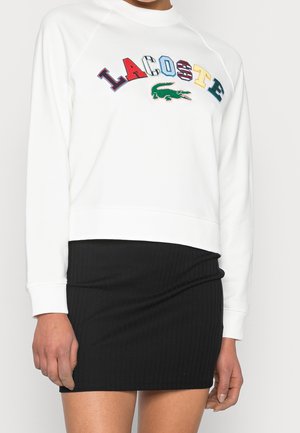 Biała bluza Lacoste z wielokolorowymi literami i zielonym logo krokodyla, noszona z czarną prążkowaną minispódnicą.