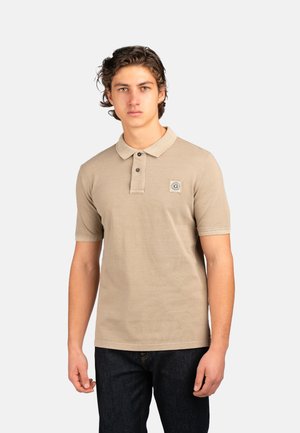 Camisa polo beige con cuello y tapeta de dos botones. Presenta una tela texturizada y un parche con logo en el lado izquierdo del pecho. Combinada con jeans oscuros.