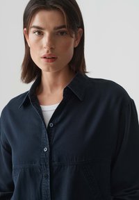 Dark denim button-up shirt featuring a spread collar, front pockets, and a relaxed fit, layered over a white ribbed top.