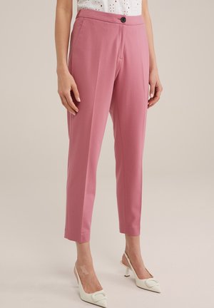 Kobieta ubrana w różowe spodnie o zwężanych nogawkach z wysokim stanem, czarnym guzikiem oraz białe sandały na obcasie typu slingback, stojąca na neutralnym tle.