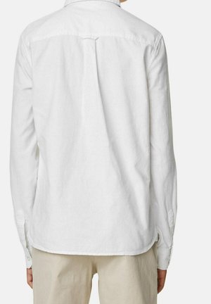 Hombre vestido con camisa blanca de manga larga abotonada y pantalones beige, mostrado de espaldas sobre un fondo liso.