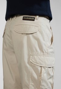 Beige cargo šortky vyrobené z lehkého materiálu, s množstvím kapes, nášivkou na pásku a viditelnými švy.