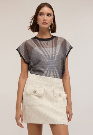 Durchsichtige schwarze Bluse mit kurzen Ärmeln, die horizontale Streifen und ein geometrisches Muster aufweist, kombiniert mit einem strukturierten cremefarbenen Minirock mit Taschen.