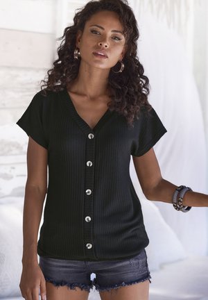 Zwart, geribde gebreide top met korte mouwen, V-hals en zilveren knopen aan de voorkant. Gecombineerd met denimshorts en meerdere armbanden.
