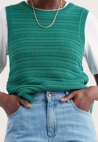 Green knitted sleeveless top featuring textured stripes, worn over a white shirt, paired with light blue denim jeans. Gold chain necklace visible.