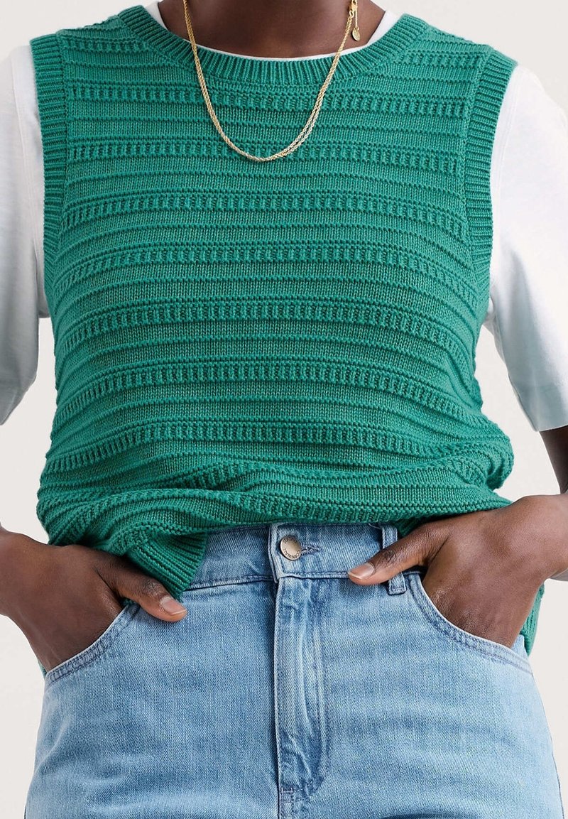 Green knitted sleeveless top featuring textured stripes, worn over a white shirt, paired with light blue denim jeans. Gold chain necklace visible.