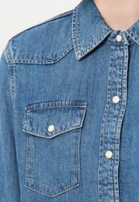 Niebieska koszula dżinsowa z wywiniętym kołnierzem, kieszenią z przodu z zapięciem na guzik oraz srebrnymi guzikami wzdłuż listwy. Miękka tekstura.