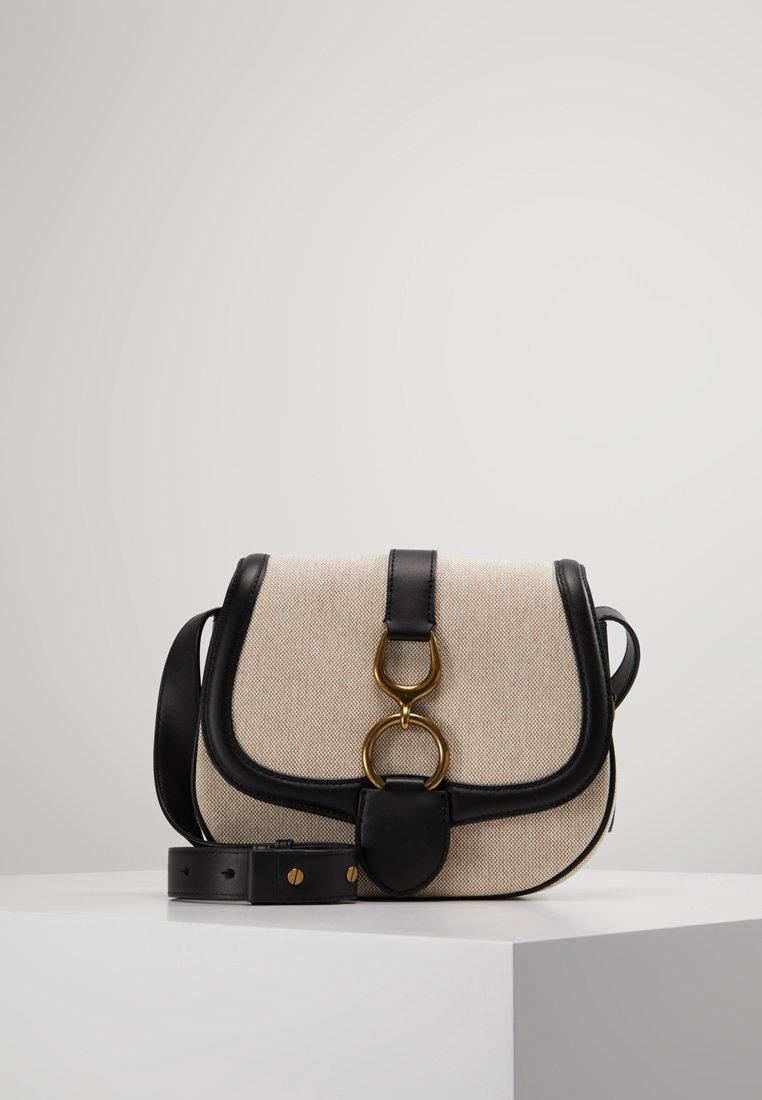 Sac bandoulière en toile beige avec des garnitures en cuir noir, fermoir circulaire doré et bandoulière en cuir noir ajustable sur un piédestal blanc.