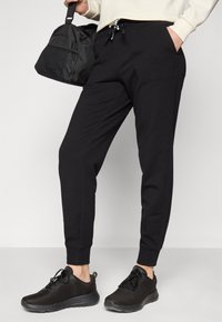 Svarte joggebukser med tapered design, elastisk midjebånd og lommer. Kombinert med svarte mesh joggesko og en svart treningsveske.