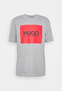 Camiseta de algodón gris con un audaz logotipo rectangular rojo con "HUGO" y "HUGO BOSS" impresos en negro. Cuello redondo simple y mangas cortas.