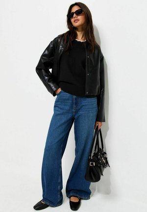 Donna con giacca di pelle nera, maglione nero, jeans blu a gamba larga, scarpe nere e occhiali da sole, che tiene una borsa nera.