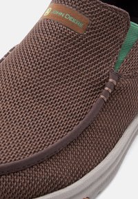 Καφέ πλεκτό παπούτσι χωρίς κορδόνια με πράσινη εσωτερική επένδυση και λευκή σόλα, με δερμάτινο λογότυπο "John Deere" στο επάνω μέρος.