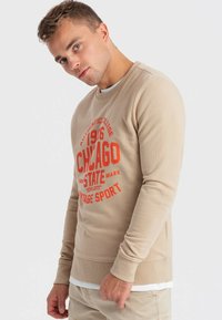 Felpa beige con texture a coste, caratterizzata da una stampa grafica audace di colore rosso che recita "1966 CHICAGO STATE VINTAGE SPORT." Collo rotondo, polsini aderenti.