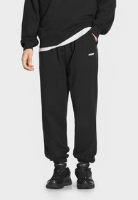 Czarne dresowe spodnie z elastycznym pasem i mankietami, z małym białym logo, zestawione z błyszczącymi czarnymi butami sportowymi.