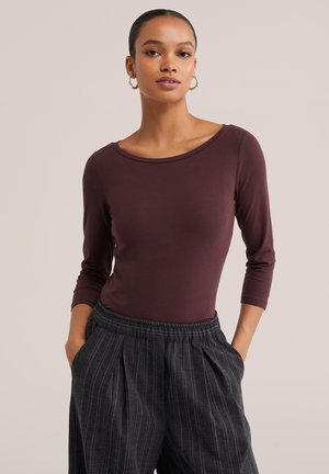 Femme aux cheveux tirés en arrière, portant des créoles en or, un haut ajusté à manches longues violet foncé, et un pantalon ample gris foncé à fines rayures, les mains dans les poches.
