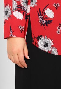 Rød blomsterbluse med sorte accenter. Har lange ærmer, en rund bund og intrikate blomsterdesigns i hvidt og pink på stoffet.