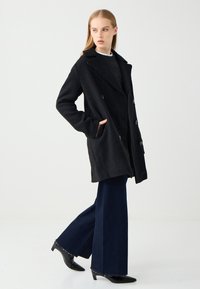 Schwarzer Wollmantel mit großen Revers und doppelt geknöpften Knöpfen, mit Seitentaschen. Kombiniert mit marineblauen weit geschnittenen Jeans und schwarzen spitzen Schuhen.