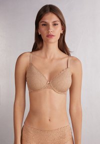 Top de encaje en tono nude, con forma triangular, tirantes ajustables y un delicado lazo en el centro.