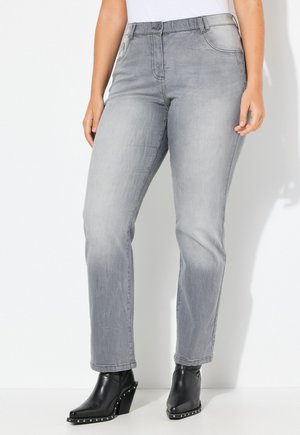 Person trägt hellgraue Jeans mit geradem Bein und schwarze, mit Nieten besetzte Stiefeletten mit Absatz, steht vor weißem Hintergrund.