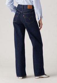Personne portant un jean droit bleu foncé et une chemise bleu clair à manches longues, debout de dos à la caméra, chaussures à talons argentés visibles.