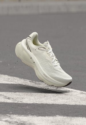 Chaussure de sport blanche en l'air au-dessus d'une route asphaltée avec des lignes blanches peintes et de petites particules de gravier visibles.