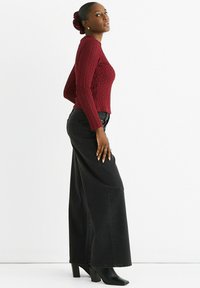 Woman in a textured red sweater, wide-leg black jeans, and black heeled boots, standing sideways against a white background.