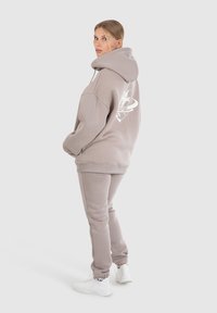 Lichtgrijs hoodie met bijpassende trainingsbroek van zacht materiaal, met een wit grafisch logo op de achterkant. Draagt witte schoenen.
