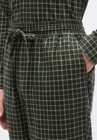 Pantalones a cuadros verdes con cintura elástica, cordón ajustable y bolsillos laterales, que presentan un patrón de cuadros en verde oscuro, beige y blanco.