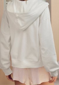 Vue arrière d'une personne portant un hoodie blanc ample à longues manches et un short rose clair sur fond beige.