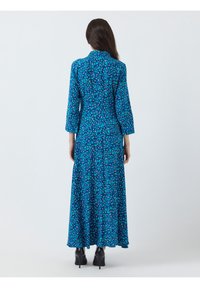 Robe longue en tissu bleu avec un motif floral. Manches longues, corsage ajusté et jupe évasée. Portée avec des chaussures à talons noirs.
