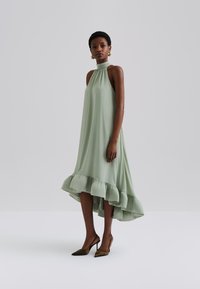 Luftig, ärmløs kjole i myk grønn stoff, med samlet halterneck og volangkant i en høy-lav design.