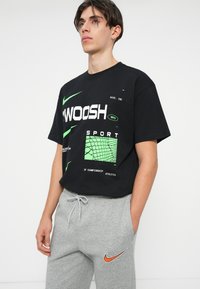 Melna kokvilnas t-krekls ar izteiksmīgu balto un zaļo "SWOOSH" tekstu. Kombinēts ar gaiši pelēkiem sporta biksēm, kurām ir oranžs Nike logotips.
