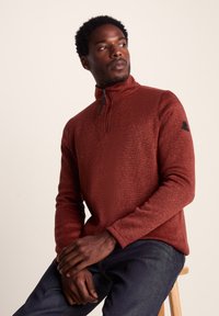 Maroon zip-up pullover with a textured finish, featuring a stand-up collar and black logo accent on the sleeve. Worn with dark jeans.