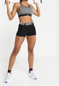 Brassière de sport grise avec bretelles noires, short de compression noir avec ceinture "PRO NIKE", et chaussures de sport blanches avec des accents roses.