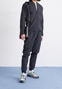 Osoba ve černé zipové bundě a kalhotách, s bílými a šedými sportovními botami, držící černou křížovou tašku, stojící na modré podlaze.